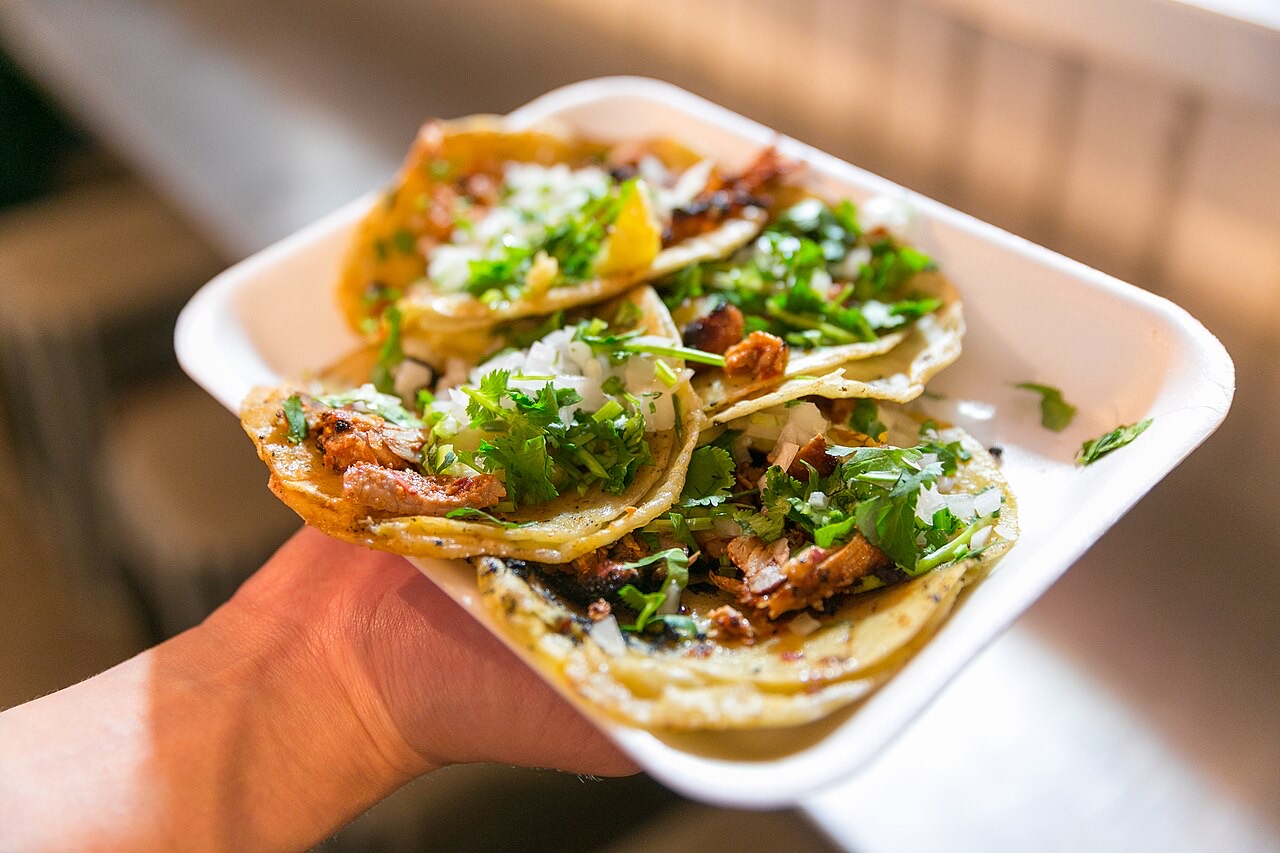 A hand holding a tray of tacos al pastor topped with cilantro and onion